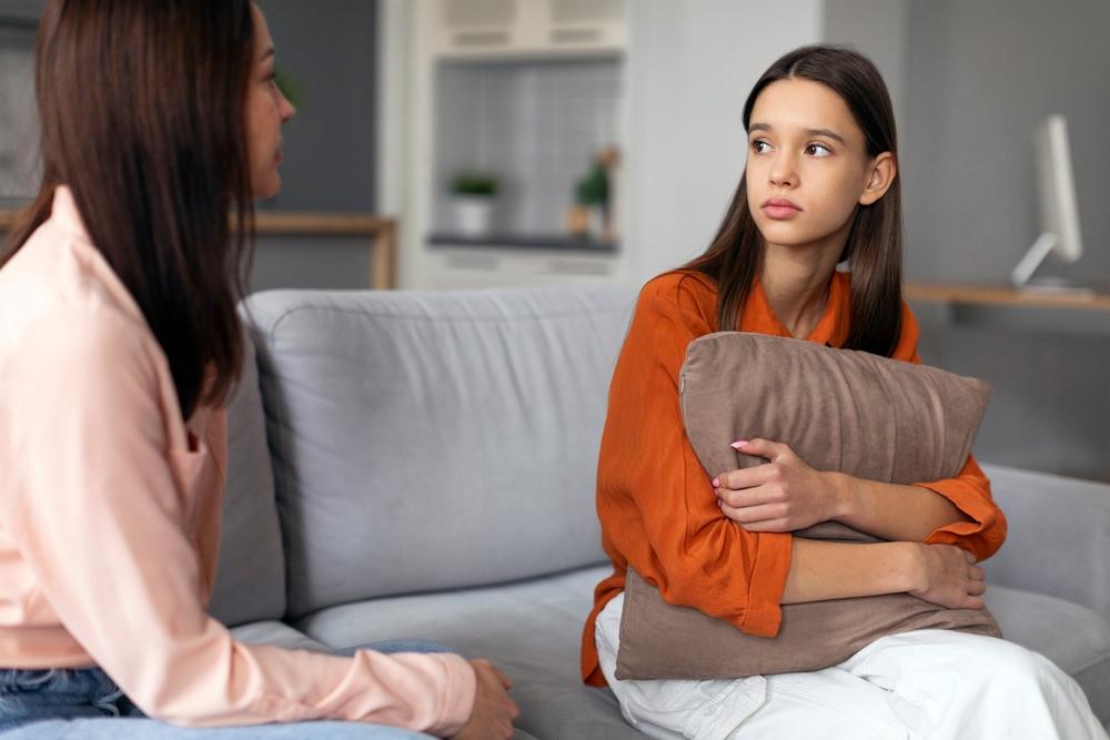 Adolescente sentada no sofá, abraçando um travesseiro, conversa com uma mulher adulta em um ambiente doméstico, representando um momento de apoio e diálogo sobre gravidez na adolescência.