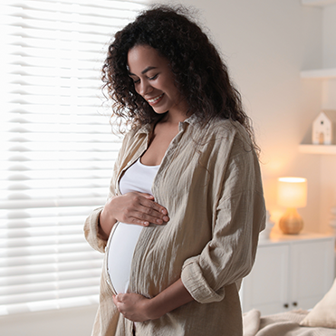 Mulher em pé em ambiente bem iluminado, com persianas ao fundo, vestindo cardigã claro sobre blusa branca, com as mãos sobre a barriga. Ao fundo, itens de decoração como prateleiras e luminárias.