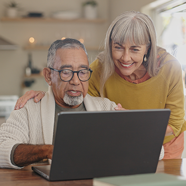 Um casal de idosos interagindo com notebook em ambiente interno iluminado por luz natural. O homem usa óculos e está sentado, enquanto a mulher apoia a mão no ombro, sugerindo colaboração, aprendizado digital e uso de tecnologia em ambiente doméstico.