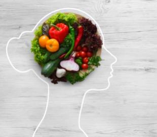 Um perfil de cabeça humana contornado por uma linha branca, com um prato de vegetais coloridos (tomates, pimentões, alface, pepino) sobre uma tábua de madeira no lugar do cérebro, simbolizando a importância da alimentação saudável para combater o colesterol.