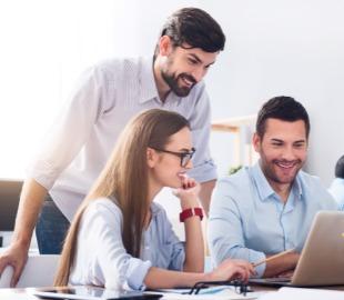 Três colegas de trabalho sorrindo e interagindo em frente a um laptop, simbolizando os benefícios de contratar um plano de saúde para colaboradores e a satisfação no ambiente de trabalho.