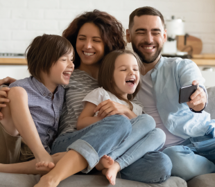 Família feliz (pai, mãe e dois filhos) sorrindo e se divertindo em um sofá, enquanto o pai segura um celular para tirar uma selfie, simbolizando a qualidade do atendimento do Centro Clínico Joinville.