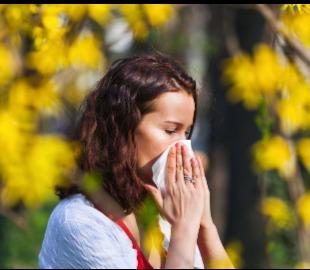Mulher com cabelos ruivos assoando o nariz em um lenço de papel, com árvores de flores amarelas desfocadas ao fundo, simbolizando alergias e doenças comuns da primavera.