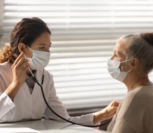 Médica de família usando máscara e estetoscópio para examinar uma paciente idosa, ambas de máscara, simbolizando o cuidado com a saúde familiar.