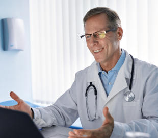 Médico de jaleco e óculos, sorrindo e gesticulando em uma teleconsulta, simbolizando um serviço de saúde completo na APS Florianópolis