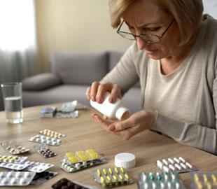 Mulher idosa com óculos derramando comprimidos de um frasco, com diversas cartelas de medicamentos sobre a mesa, simbolizando os riscos da automedicação.