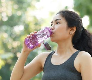 Mulher usando regata de ginástica, bebendo água de uma garrafa roxa em ambiente externo, simbolizando a importância da hidratação para a saúde.