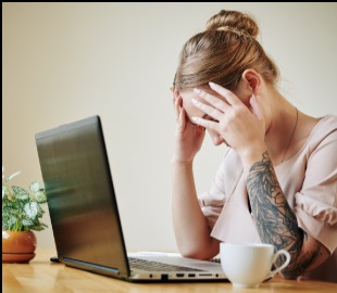 Mulher sentada em frente a um laptop, com as mãos na cabeça e expressão de desespero, simbolizando o esgotamento e a Síndrome de Burnout.