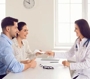 Casal sorridente em consulta com uma médica de jaleco e estetoscópio, que entrega documentos, simbolizando a escolha de um plano de saúde.