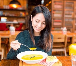 Mulher sorridente saboreando uma tigela de sopa quente em um ambiente acolhedor, simbolizando a preparação de sopas saudáveis no inverno.