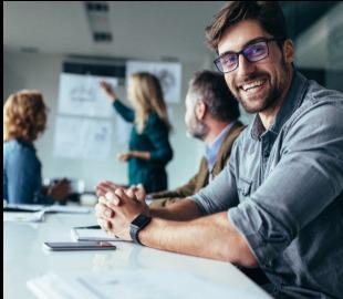 Homem sorridente de óculos em primeiro plano durante uma reunião de negócios, com colegas de trabalho ao fundo em um escritório, simbolizando os benefícios de oferecer plano de saúde em empresas.