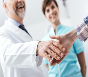 Profissional de saúde de jaleco branco apertando a mão de um paciente, com outro profissional sorrindo ao fundo, simbolizando a relação e os acordos em planos de saúde com coparticipação.