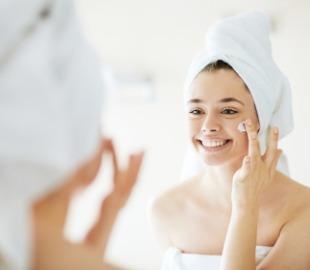 Mulher sorridente com toalha na cabeça e no corpo, aplicando creme no rosto em frente a um espelho, ilustrando uma rotina de skincare para cuidar da pele.
