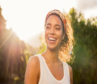 Mulher jovem sorridente aproveitando o sol em um ambiente externo, simbolizando o cuidado com a saúde no verão.