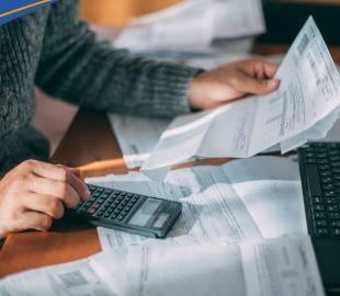 Mãos de uma pessoa segurando uma fatura e usando uma calculadora sobre uma mesa cheia de papéis, simbolizando o gerenciamento de finanças e a alteração no vencimento de faturas.