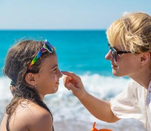 Mulher aplicando protetor solar no nariz de uma criança na praia, simbolizando a prevenção do câncer de pele na campanha Dezembro Laranja.