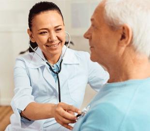 Médica sorridente examinando um paciente idoso com um estetoscópio, simbolizando a consulta médica e o tema da andropausa para o Dia do Homem.