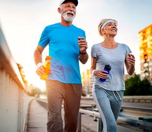 Casal de idosos sorrindo e correndo ao ar livre, segurando garrafas de água, simbolizando hábitos saudáveis e longevidade para o Dia Nacional da Saúde.