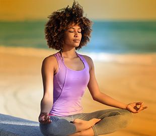 Mulher de cabelo cacheado em pose de lótus na praia, com os olhos fechados e uma expressão serena, simbolizando a meditação para iniciantes.