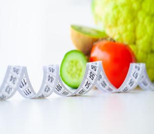Fita métrica enrolada com frutas e vegetais (kiwi, pepino, tomate e brócolis) ao fundo, simbolizando a dieta keto e a perda de peso.
