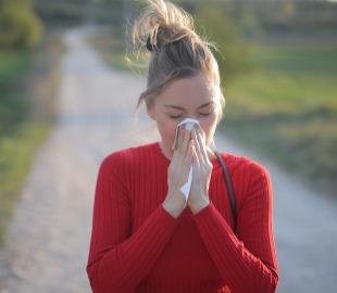 Mulher assoando o nariz em um lenço de papel, com um caminho ao ar livre desfocado ao fundo, simbolizando os sintomas de doenças comuns do outono, como resfriados ou alergias.