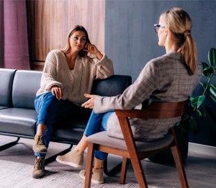 Mulher sentada em um sofá, olhando para sua terapeuta que está sentada de frente para ela, ambas conversando em uma sessão de terapia, simbolizando o apoio para lidar com problemas e a importância da terapia.