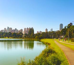 Pista de caminhada/corrida às margens de um lago com a paisagem urbana de Curitiba ao fundo, sob um céu azul, simbolizando locais para se exercitar na cidade.