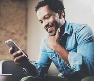 Homem negro sorridente, usando uma camisa jeans, olhando para o celular, simbolizando a facilidade de acessar um extrato de utilização online.