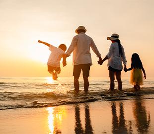 Família (pai, mãe e dois filhos) de mãos dadas, de costas para a câmera, caminhando na praia ao pôr do sol, simbolizando férias e a importância da saúde preventiva em viagens.
