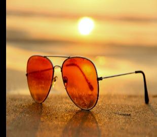 Óculos de sol com lentes alaranjadas refletindo o sol poente em uma praia, simbolizando a proteção contra o sol e os riscos de insolação e golpe de calor.
