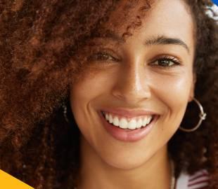Mulher de cabelo cacheado sorrindo e mostrando dentes saudáveis, simbolizando uma nova experiência e cuidado completo com a saúde bucal e o sorriso.