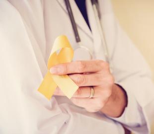 Mão de um médico de jaleco branco e estetoscópio segurando uma fita amarela de conscientização, simbolizando a prevenção de hepatites virais.