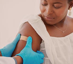 Profissional de saúde com luvas azuis aplicando um curativo no braço de uma mulher após uma vacina, simbolizando a prevenção de doenças como a coqueluche.