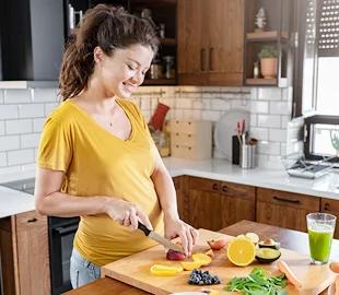 Mulher grávida sorrindo na cozinha, cortando frutas e vegetais frescos, simbolizando os cuidados com a alimentação saudável durante a gravidez.