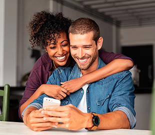 Casal sorridente e feliz abraçado, olhando para o celular, simbolizando a facilidade de usar um aplicativo de saúde como o Clinipam para cuidar da saúde.