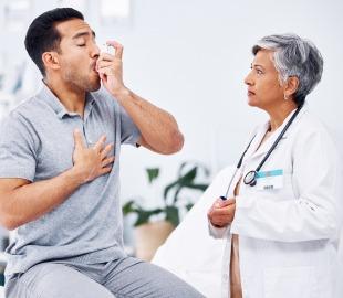 Homem usando um inalador enquanto uma médica de cabelos grisalhos e estetoscópio o observa com preocupação, ilustrando a asma grave e seus riscos.