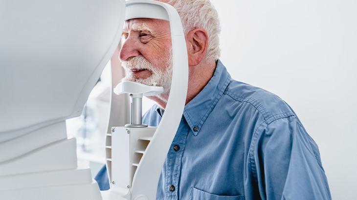 Homem idoso com barba branca sendo examinado em um aparelho oftalmológico, ilustrando um exame de visão para condições como astigmatismo.