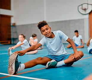 Criança sorridente praticando alongamento em uma quadra de esportes com outras crianças ao fundo, ilustrando atividades físicas infantis.