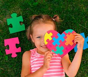 Criança sorridente deitada na grama, segurando um coração feito de peças de quebra-cabeça coloridas, com peças soltas ao lado, simbolizando a conscientização e o diagnóstico do autismo.