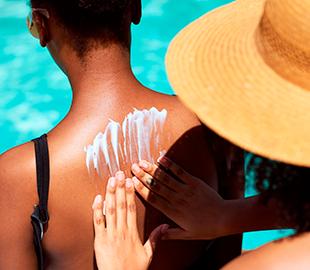 Pessoa aplicando protetor solar nas costas de outra pessoa, ambas em um ambiente de piscina ou praia, simbolizando a prevenção do câncer de pele no Dezembro Laranja.