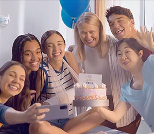 Grupo de jovens e uma mulher mais velha sorrindo, fazendo uma selfie em um quarto de hospital com um bolo de aniversário, simbolizando a campanha institucional Sinal de Vida da Hapvida.
