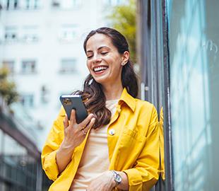 Mulher sorrindo, usando uma camisa amarela e olhando para seu celular, simbolizando a praticidade de atualizar dados em um portal ou aplicativo.