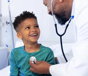 Médico de jaleco examinando um menino sorridente com um estetoscópio, simbolizando a inclusão de dependentes em um plano de saúde e o cuidado pediátrico.
