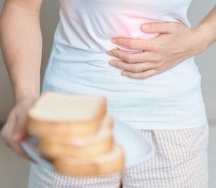 Pessoa com dor abdominal segura o estômago enquanto oferece pão, ilustrando a relação entre glúten e os sintomas da doença celíaca.