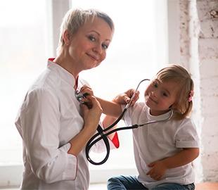Médica sorridente de cabelos curtos observando uma criança com síndrome de Down que brinca com um estetoscópio, simbolizando o cuidado e a compreensão de doenças genéticas.