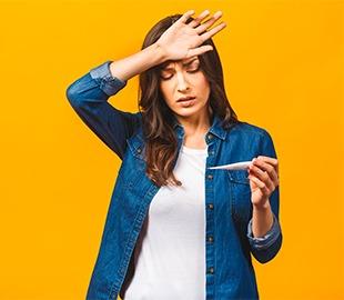 Mulher com a mão na testa, parecendo indisposta, segurando um termômetro que indica febre, simbolizando sintomas de doenças tropicais.