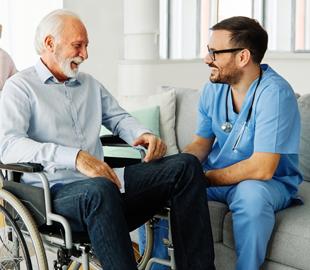 Médico sorridente de óculos sentado em um sofá conversando com um paciente idoso em uma cadeira de rodas, simbolizando o cuidado e a conscientização sobre a Esclerose Múltipla no Agosto Laranja.