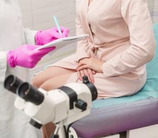 Consulta ginecológica onde uma médica de luvas rosas anota informações em uma prancheta, enquanto uma paciente senta na maca. Em primeiro plano, um colposcópio, simbolizando exames ginecológicos e saúde da mulher.