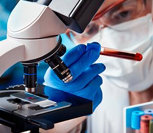 Cientista ou hematologista com luvas azuis e máscara, observando uma amostra em um microscópio e segurando um tubo de ensaio com sangue, simbolizando a função do hematologista.