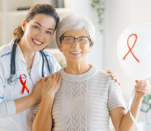 Médica sorridente e paciente idosa, ambas com uma fita vermelha de conscientização, segurando um balão com a mesma fita, simbolizando o Maio Vermelho e a conscientização sobre doenças cardiovasculares.